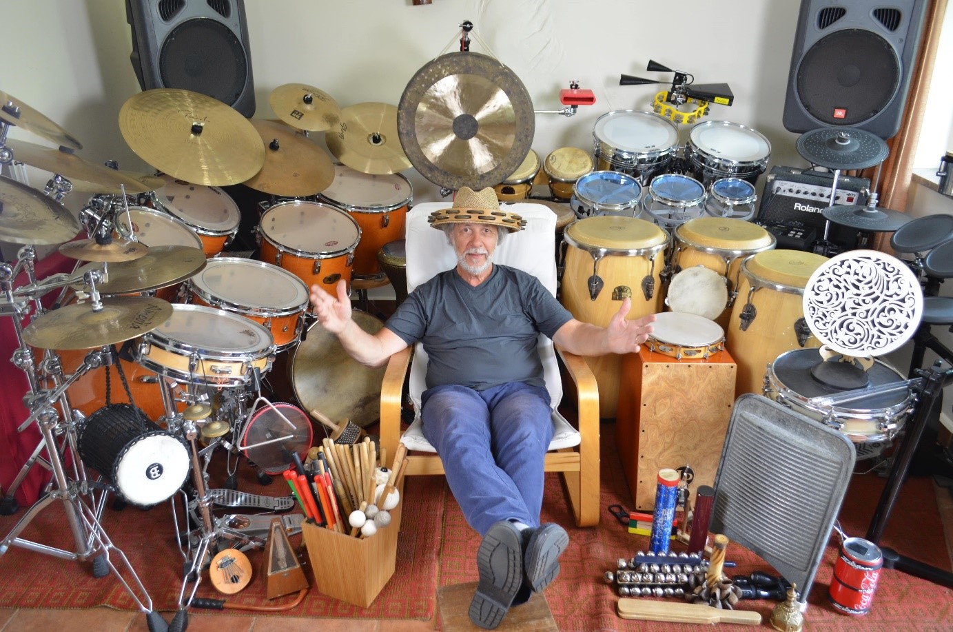 Obraz przedstawiający rozłożone instrumenty na całej szerokości pokoju: zestawy perkusyjne, kongi, talerze, rototomy, cajon i wiele innych.

Na środku jest prowadzący z rozłożonymi rękami i tamburynem na głowie.